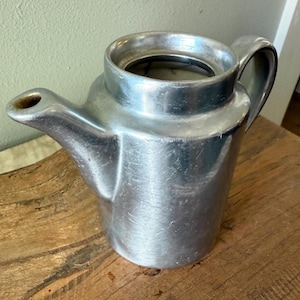 Puede incluir: Una jarra de aluminio cepillado con boquilla y asa. La jarra tiene un cuerpo cilíndrico y una amplia abertura en la parte superior. La boquilla es pequeña e inclinada. El asa es curva y está unida al lateral de la jarra.