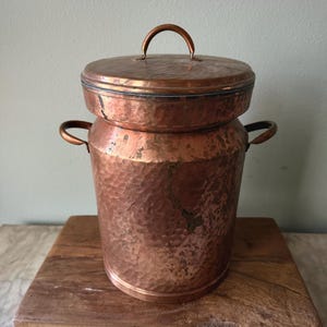 May include: A vintage copper container with a hammered texture, featuring a lid with a handle and two side handles. The container is cylindrical and sits on a wooden surface. The copper has a warm, reddish-brown hue.