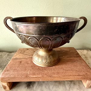 May include: A silver-toned metal bowl with two curved handles. The bowl sits on a decorative pedestal base with a scalloped design. The bowl is placed on a wooden stand.