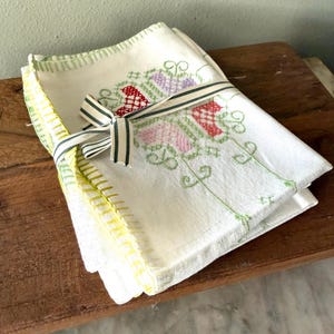 Könnte beinhalten: Ein Stapel weißer Leinen-Servietten mit gestickten Blumenmustern in Rot, Rosa und Lila, gebunden mit einem gestreiften Band. Die Ränder sind mit grünen und gelben Nähten versehen. Die Servietten liegen auf einer Holzoberfläche.