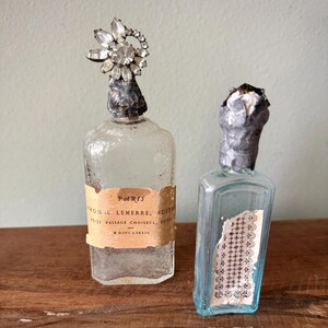 May include: Two antique perfume bottles. One is clear with a crystal flower stopper and a label that reads "Paris". The other is a small, light blue bottle with a label and a silver-colored stopper. Both bottles are on a wooden surface.