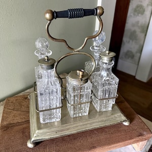 May include: A condiment set featuring four clear glass bottles with square bases and metal caps, held in a brass-toned metal caddy. The caddy includes a central arched handle with a black textured grip and rests on a rectangular metal base.
