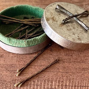 May include: An open, round, antique metal container with a green velvet interior filled with brown bobby pins. Two bobby pins rest outside the container on a wooden surface. The lid is silver and has two bobby pins crossed on top.