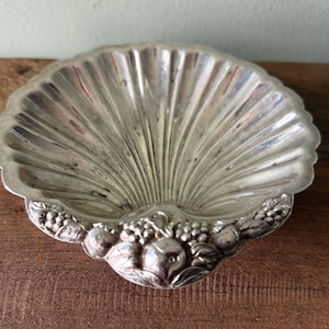 Puede incluir: Un plato plateado en forma de concha con un borde festoneado. El plato tiene un interior acanalado y está decorado con un diseño detallado de frutas y hojas en la base. El plato es probablemente de metal y se exhibe sobre una superficie de madera.