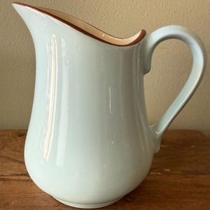 Puede incluir: Una jarra de cerámica azul claro con un asa curva y un pico. El borde está adornado con una banda marrón. La jarra se utiliza probablemente para servir bebidas y tiene un acabado brillante.