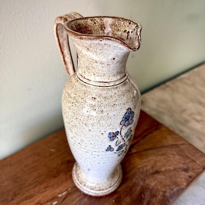 May include: A speckled ceramic pitcher with a handle and a spout. The pitcher is primarily off-white with brown speckles and a brown rim. Blue floral designs decorate the body of the pitcher. The pitcher is on a wooden surface.
