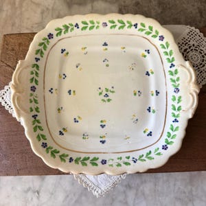 May include: A square, off-white ceramic serving platter with a scalloped edge and handles. The platter is decorated with green and blue floral patterns around the border and scattered across the surface. A gold-colored line accents the edge.