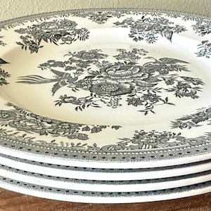 May include: A stack of four white ceramic plates with a gray floral design. The plates have a detailed border and a central floral motif. The design includes roses, other flowers, and a bird. The plates are stacked on top of each other.