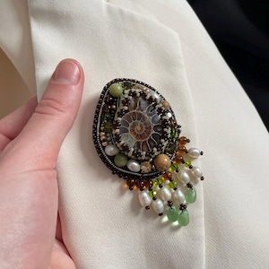 Puede incluir: Broche ovalado con diseño de fósil, con un centro en espiral y rodeado de cuentas en tonos marrón, verde y crema. El broche tiene un fleco de perlas blancas y cuentas verdes en forma de lágrima. El broche está sujeto a una blazer color crema.