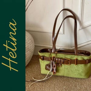 May include: A lime green handbag with a dark brown belt-style strap across the front, featuring a gold-tone buckle, two thin brown shoulder straps, and a decorative charm with white and brown accents. The word Hetina is written in yellow.