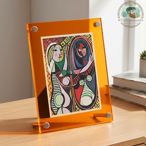 Peut inclure: Un cadre photo en acrylique orange avec une impression d'art abstrait colorée. Le cadre est maintenu par des vis argentées et possède un petit support. L'impression d'art présente un visage de femme et d'autres formes abstraites. Le cadre est sur une surface en bois.