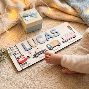 May include: A rectangular wooden name puzzle featuring the name LUCAS in large blue letters, accompanied by wooden cutouts of a helicopter, police car, cement truck, and ambulance, each labeled with its respective name in black text.