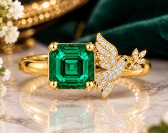 May include: A gold ring featuring a square-cut green gemstone held by four prongs. Attached to the side of the setting is a gold bird motif encrusted with small white stones, with the bird holding a small branch in its beak.