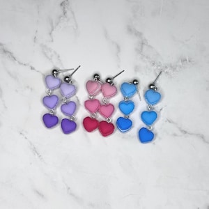 Puede incluir: Tres pares de pendientes colgantes con dijes en forma de corazón en tonos morado, rosa y azul. Cada pendiente tiene tres corazones graduados suspendidos de un poste plateado. Los pendientes se exhiben sobre una superficie de mármol blanco.