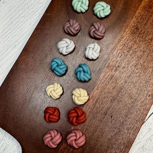 Peut inclure: Une collection de boucles d'oreilles en forme de nœuds, aux couleurs variées : vert menthe, mauve, blanc, sarcelle, jaune, rouge et rose. Les boucles d'oreilles sont disposées sur une planche à découper en bois brun foncé, mettant en valeur leur design unique et leurs couleurs vives.