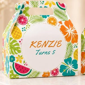 May include: Two gable-style paper favor boxes featuring a vibrant tropical pattern of hibiscus flowers, palm leaves, pineapples, and watermelon slices. The left box reads Kenzie Turns 5, and the right box reads Thank you! Love, Kenzie in colorful text.