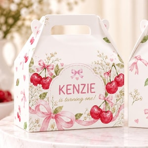 May include: Two white gable boxes featuring a watercolour print of red cherries, pink ribbons, and small white flowers. The left box displays the text "KENZIE is turning one!" in pink lettering. Both boxes have a handle at the top and a square base.