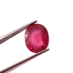 Puede incluir: Un rubí de forma ovalada, sujeto por unas pinzas plateadas sobre un fondo blanco. El rubí exhibe un color rojo intenso con ligeras variaciones tonales. Las pinzas permiten una vista detallada de las facetas y la superficie de la gema.