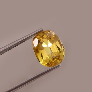 May include: A faceted, oval-shaped yellow gemstone held by metal tweezers. The gem has a brilliant, reflective surface, showcasing a range of yellow hues. The background is a neutral, light brown.