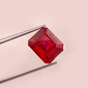 Puede incluir: Un rubí tallado en forma cuadrada, sostenido por unas pinzas plateadas. El rubí es de un color rojo intenso y saturado, con facetas internas visibles. El fondo es de un blanco suave.