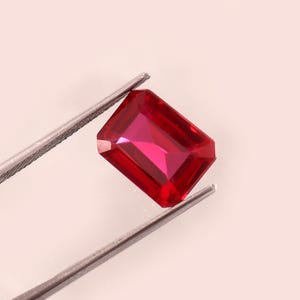 May include: A rectangular-cut ruby gemstone, held by silver tweezers. The ruby has a deep red colour and reflects light from its facets. The gemstone is the main focus, highlighting its clarity and brilliance.