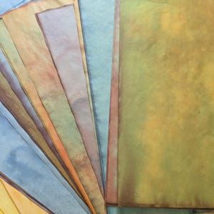 May include: A collection of rectangular paper sheets arranged in a fan shape, featuring a variety of mottled, stained, and gradient colors including shades of yellow, orange, blue, green, and brown with visible textured, aged, and weathered effects.