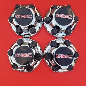 May include: Four chrome-finished GMC center caps. Each cap features a black center with the red GMC logo and is surrounded by a chrome ring and black lug nut covers. The caps are arranged on a red background.