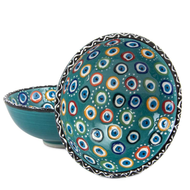 May include: Two teal ceramic bowls featuring a pattern of blue, orange, and white eye designs. The rims are decorated with a black and white pattern. The bowls are set against a white background.