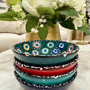 May include: A stack of four ceramic bowls in shades of teal, red, and navy blue. The top bowl is teal with a pattern of blue and white eye designs. Each bowl has a white, wavy border. A bouquet of white flowers sits in the background.