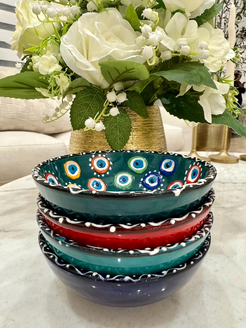 May include: A stack of four ceramic bowls in shades of teal, red, and navy blue. The top bowl is teal with a pattern of blue and white eye designs. Each bowl has a white, wavy border. A bouquet of white flowers sits in the background.