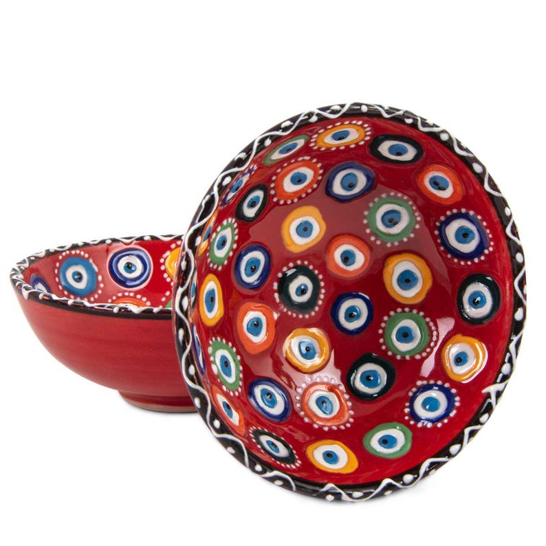 May include: Two red ceramic bowls. The larger bowl is decorated with a pattern of colourful eye-shaped designs. The smaller bowl has a similar design around the rim. The rims of both bowls are black and white.
