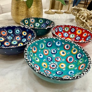May include: Four ceramic bowls with a decorative eye pattern in various colours: teal, navy, green, and red. Each bowl has a scalloped edge and is adorned with blue, white, yellow, and orange eye motifs. The bowls are arranged on a marble surface.