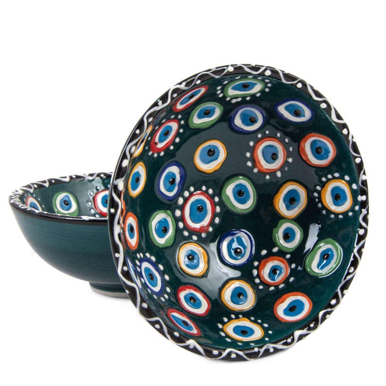 May include: Two ceramic bowls with a dark teal base colour. The bowls are decorated with a pattern of blue, white, red, and yellow eye-shaped designs. The rim of the larger bowl has a black and white decorative border.