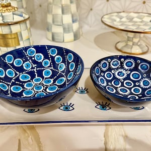 May include: Two blue ceramic bowls with white and turquoise eye designs sit on a rectangular tray with matching eye motifs. The bowls are part of a decorative set, with other matching items in the background.