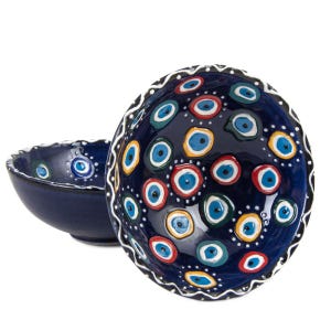 May include: Two dark blue ceramic bowls. The larger bowl is decorated with numerous colourful eye-shaped designs in blue, red, yellow, and green. The rim of the larger bowl has a white, wavy pattern. The smaller bowl is plain dark blue.