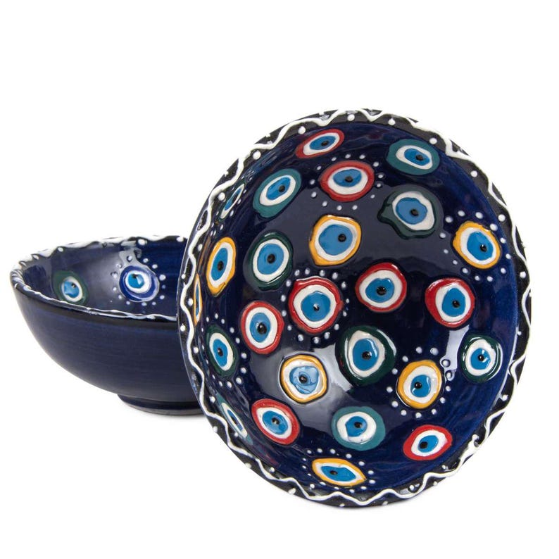 May include: Two dark blue ceramic bowls. The larger bowl is decorated with numerous colourful eye-shaped designs in blue, red, yellow, and green. The rim of the larger bowl has a white, wavy pattern. The smaller bowl is plain dark blue.