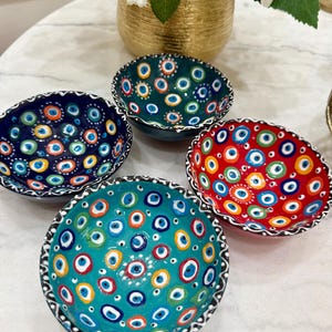 May include: Four ceramic bowls with an eye pattern in teal, red, dark blue, and green. Each bowl has a unique colour scheme. The rims are detailed with a black and white pattern. These decorative bowls would make a great addition to any home.