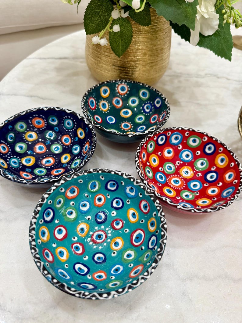 May include: Four ceramic bowls with an eye pattern in teal, red, dark blue, and green. Each bowl has a unique colour scheme. The rims are detailed with a black and white pattern. These decorative bowls would make a great addition to any home.