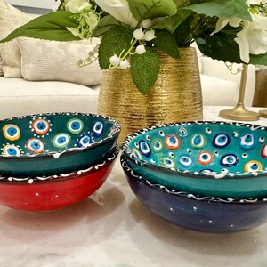 May include: A set of decorative ceramic bowls in teal, red, and navy blue. The bowls have a white and black patterned rim and are decorated with an eye-shaped design in various colours. A gold vase with flowers is in the background. The bowls are likely made in Turkey.