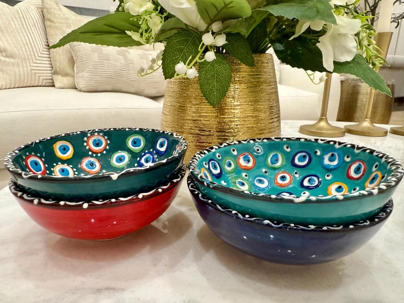 May include: A set of decorative ceramic bowls in teal, red, and navy blue. The bowls have a white and black patterned rim and are decorated with an eye-shaped design in various colours. A gold vase with flowers is in the background. The bowls are likely made in Turkey.