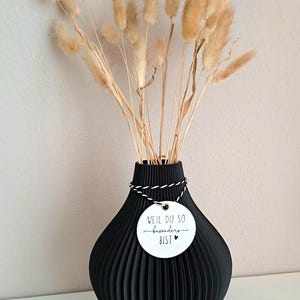 May include: A black, ribbed vase holds dried, tan-colored bunny tail grass. A white tag with black text and a heart is tied around the vase's neck. The text on the tag reads "WEIL DU SO besonders BIST".