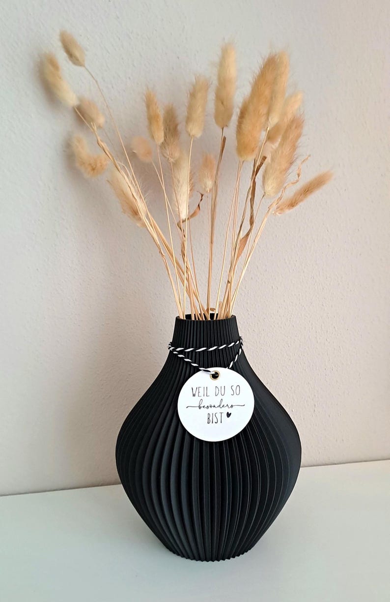 May include: A black, ribbed vase holds dried, tan-colored bunny tail grass. A white tag with black text and a heart is tied around the vase's neck. The text on the tag reads "WEIL DU SO besonders BIST".