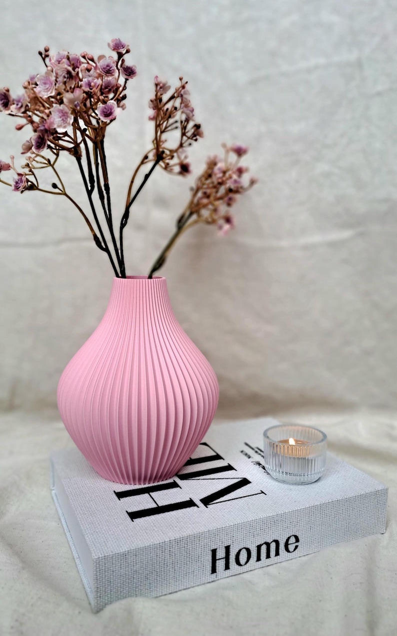 May include: A pink ribbed vase holds dried flowers, set on a book with the word "Home" visible. A small glass candle holder with a lit candle sits nearby. The arrangement is on a white surface.