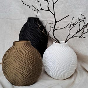 Könnte beinhalten: Drei dekorative Vasen in einer neutralen Farbpalette. Eine braune Vase, eine schwarze Vase und eine weiße Vase, jede mit einer strukturierten, geriffelten Oberfläche. Die weiße Vase enthält dunkle, zweigartige Äste, die der Anordnung ein natürliches Element hinzufügen.