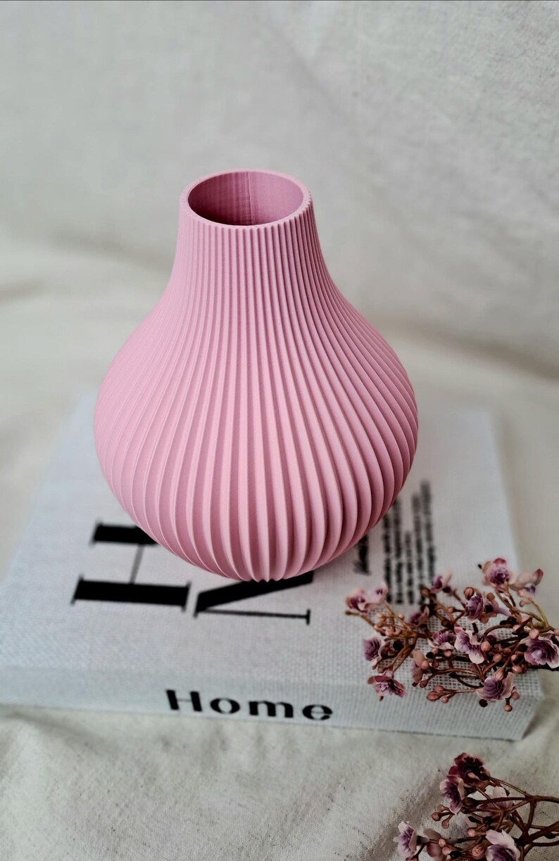 May include: A pink vase with a ribbed design. The vase is sitting on a book with the word "Home" visible. Dried flowers are next to the book. The vase is approximately 6 inches tall.