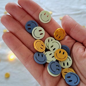 Puede incluir: Un puñado de botones de caritas sonrientes de colores en tonos amarillos, azules y verdes. Los botones son redondos con un diseño sencillo de cara sonriente. Algunos botones están esparcidos sobre una superficie blanca.