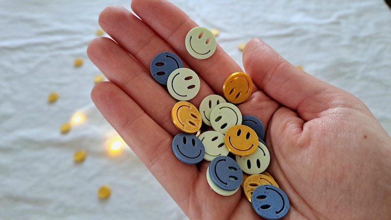 May include: A collection of smiley face buttons in various colours, including yellow, blue, and green. The buttons are circular with a simple smiling face design. Some buttons are scattered on a white surface.