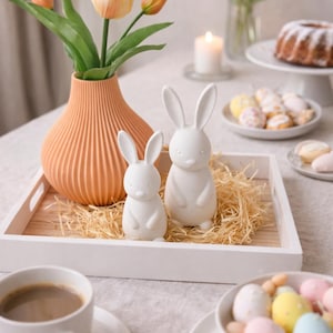 May include: A white wooden tray holds two white ceramic bunny figurines on a bed of straw. A peach-colored vase with tulips sits nearby. The scene is set for Easter, with a cup of coffee and decorated eggs.