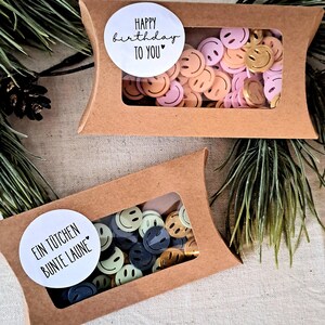 May include: Two small, brown cardboard boxes with clear windows, filled with colourful smiley face confetti. One box has a sticker that says "Happy Birthday to You." The other says "Ein T&uuml;tchen Bunte Laune."