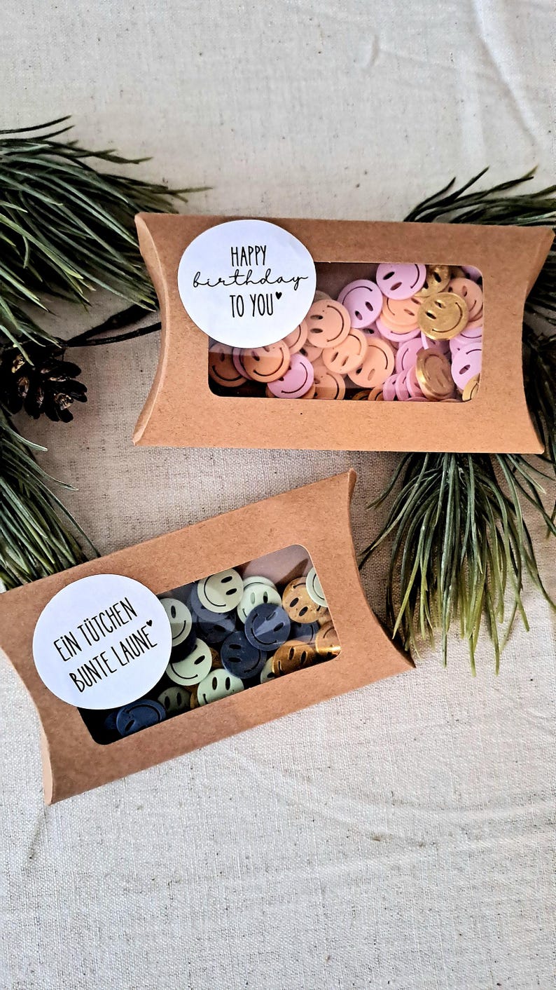 May include: Two small, brown cardboard boxes with clear windows, filled with colourful smiley face confetti. One box has a sticker that says "Happy Birthday to You." The other says "Ein T&uuml;tchen Bunte Laune."
